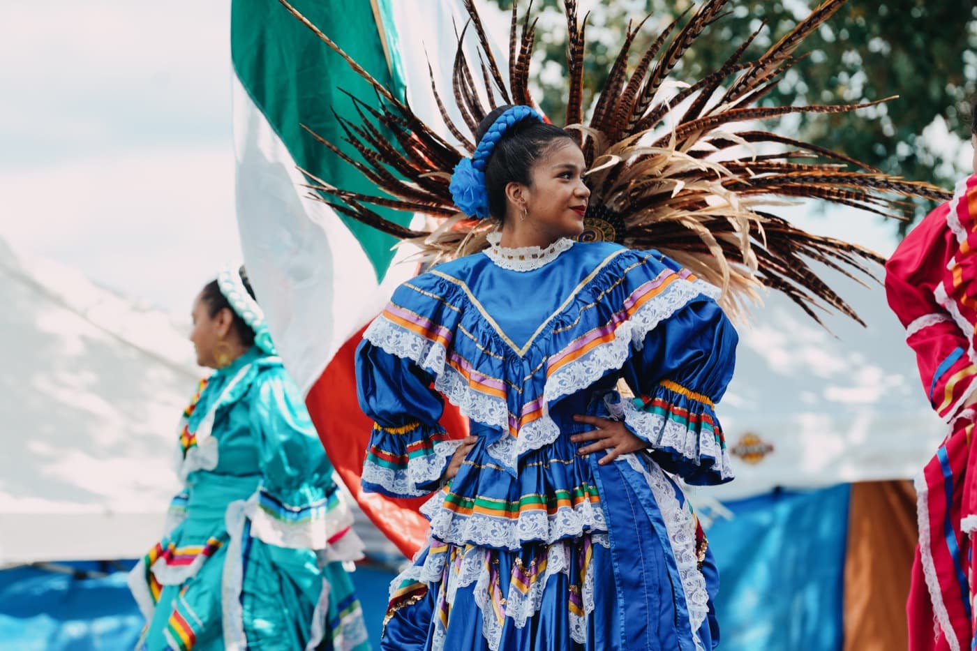 A Ballet Folklórico dancer in a bright blue ruffled dress and a feathered headdress, performing at the Madison Good Neighbor Gathering with a Mexican flag behind her