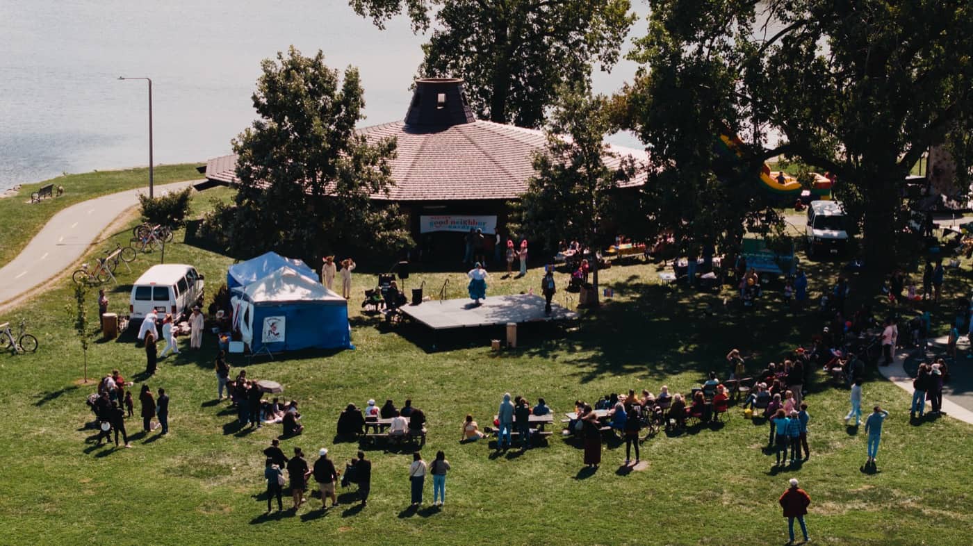 The Madison Good Neighbor Gathering seen from above: a stage beside the Brittingham Park shelter, families spread across the lawn, an inflatable slide under the trees, and Monona Bay behind