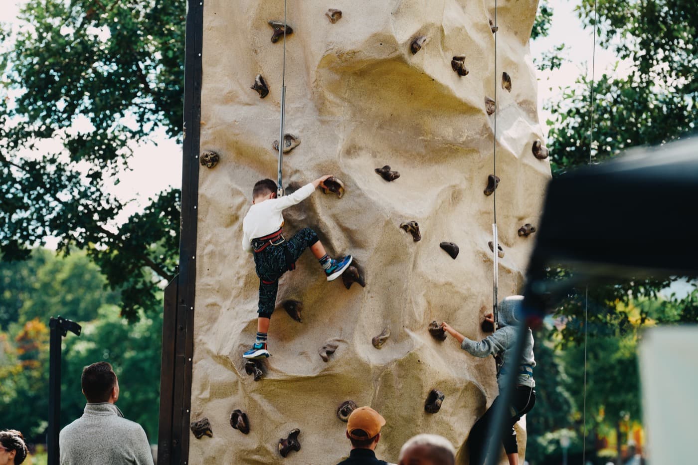 A child in a harness partway up the portable rock climbing wall at the Madison Good Neighbor Gathering, with another climber starting up beside them