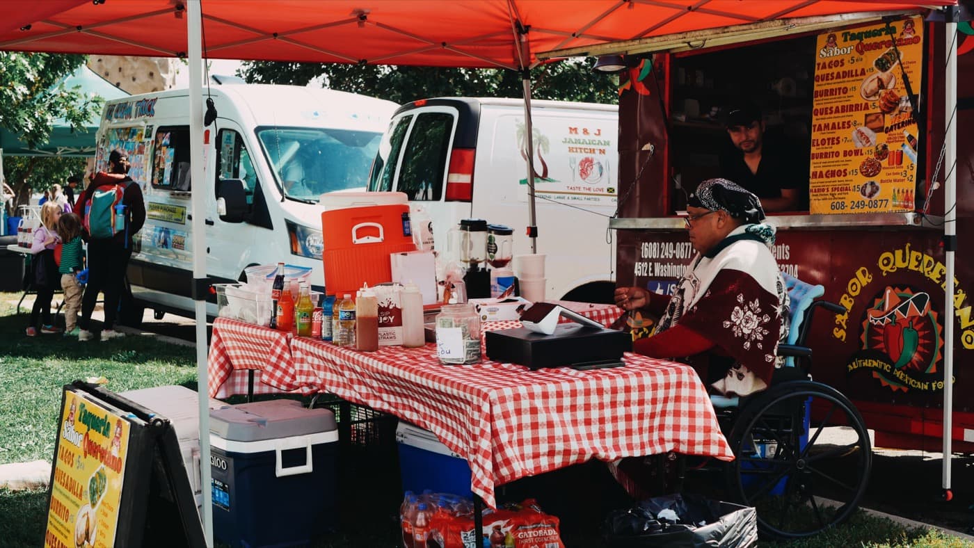 Food carts at the Madison Good Neighbor Gathering, including a taqueria and a Jamaican kitchen, with a vendor at a red gingham table under a canopy