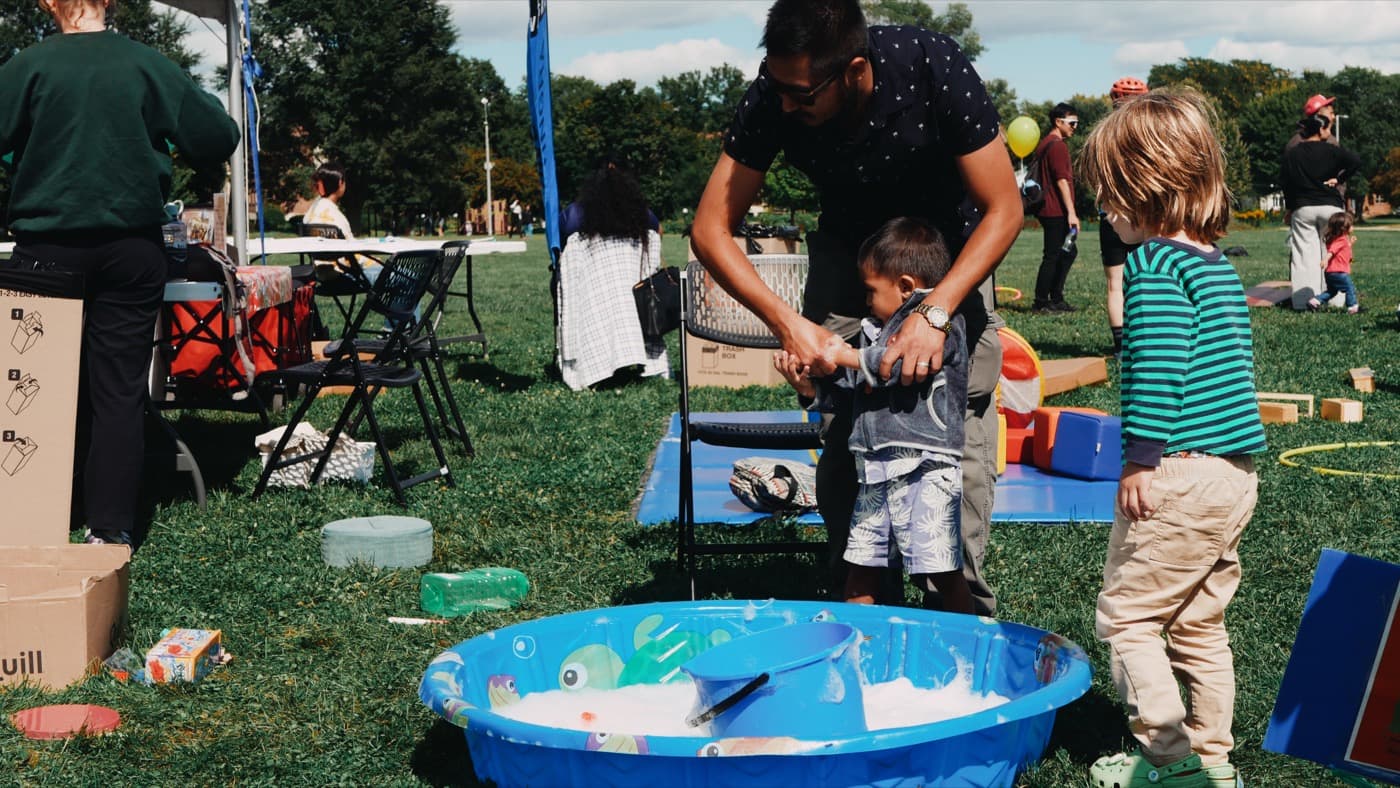 A dad helping a small boy at a bubble and water play table on the grass at the Madison Good Neighbor Gathering, with other children waiting their turn