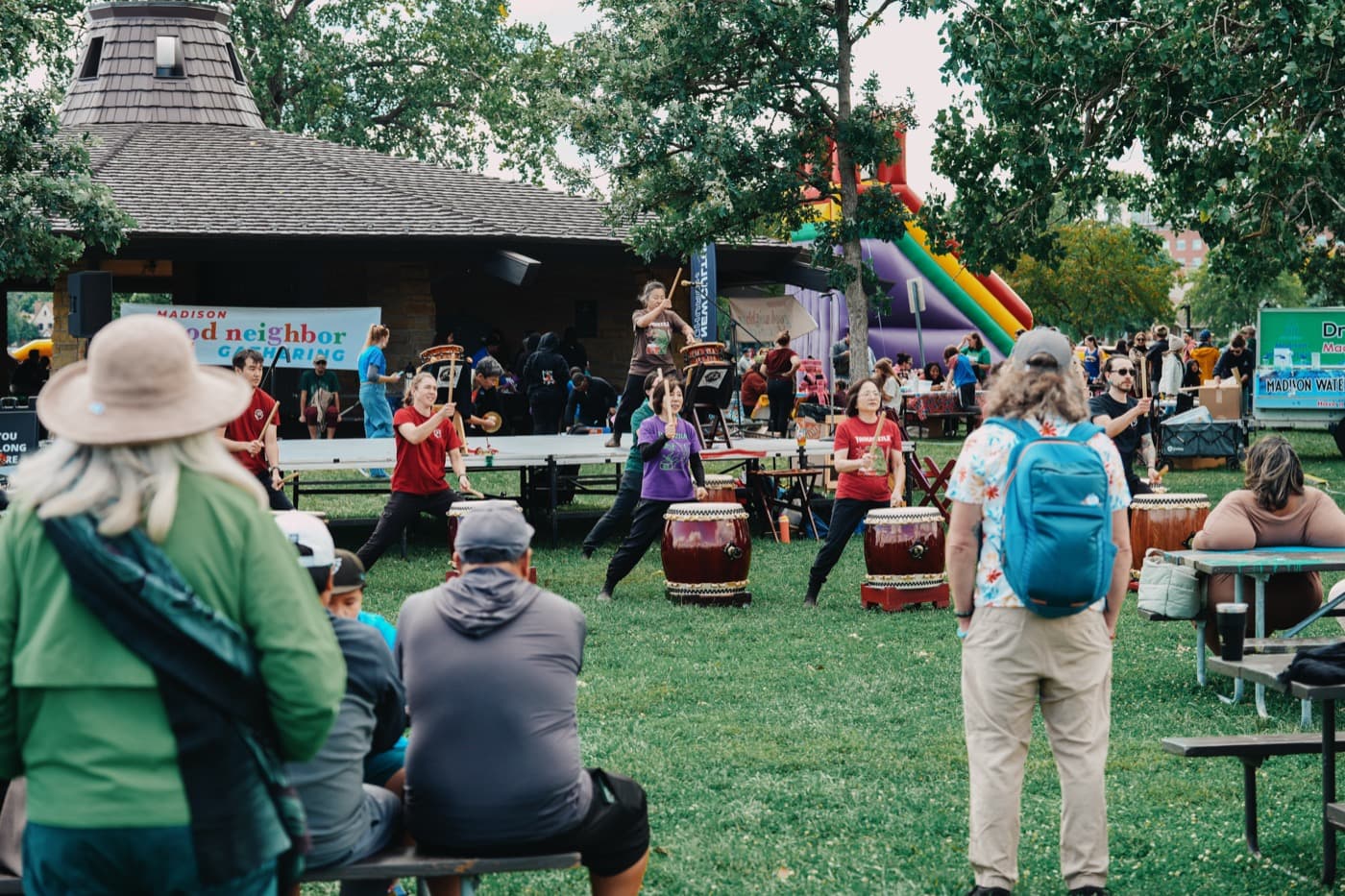 A taiko drumming ensemble performing on the lawn in front of the Brittingham Park shelter at the Madison Good Neighbor Gathering, with the festival banner behind them and neighbors watching from picnic tables