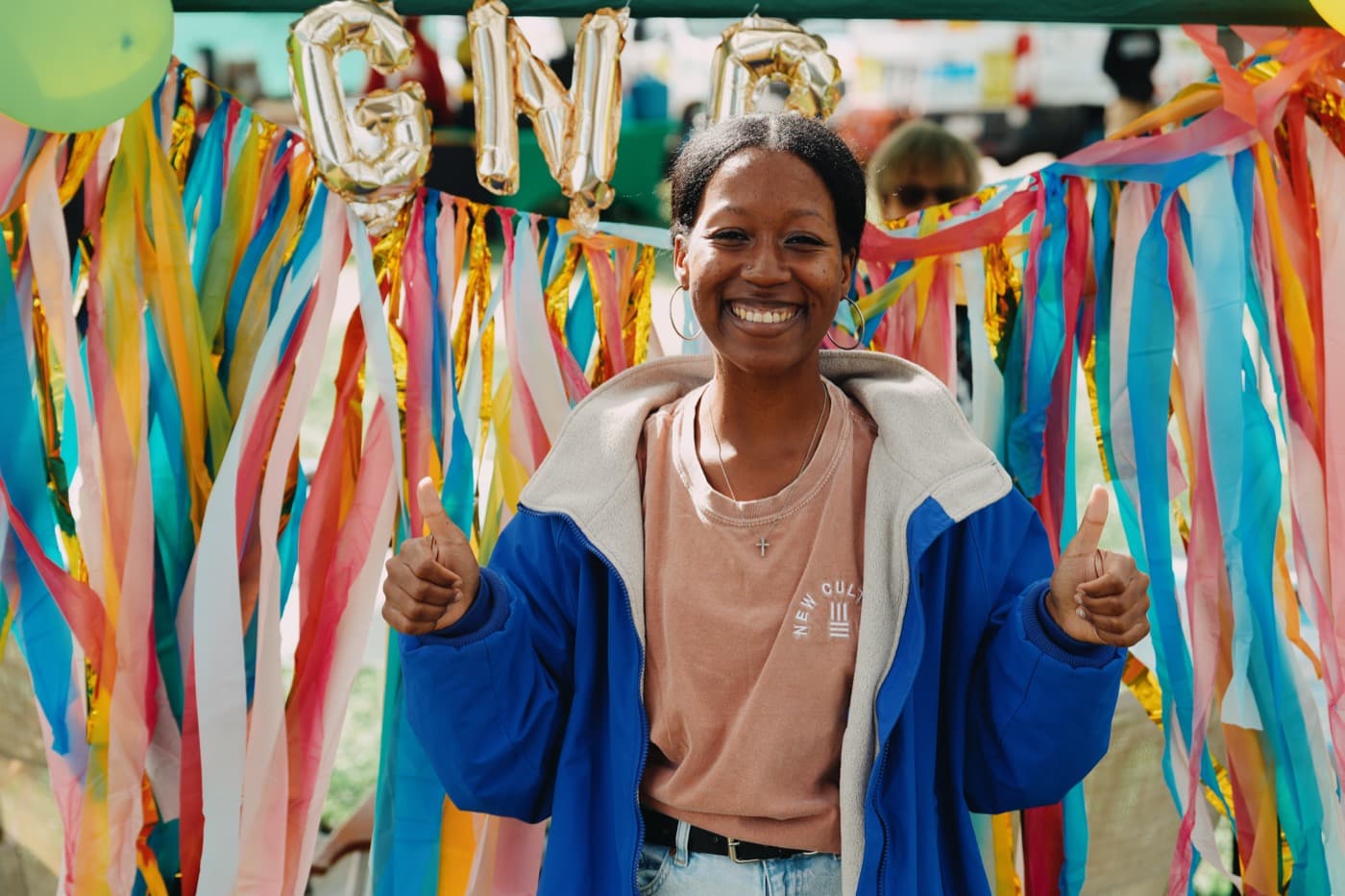 A New Culture Church volunteer in a New Culture sweatshirt giving two thumbs up in front of the colorful streamer backdrop and GNG balloons at the free family portrait booth