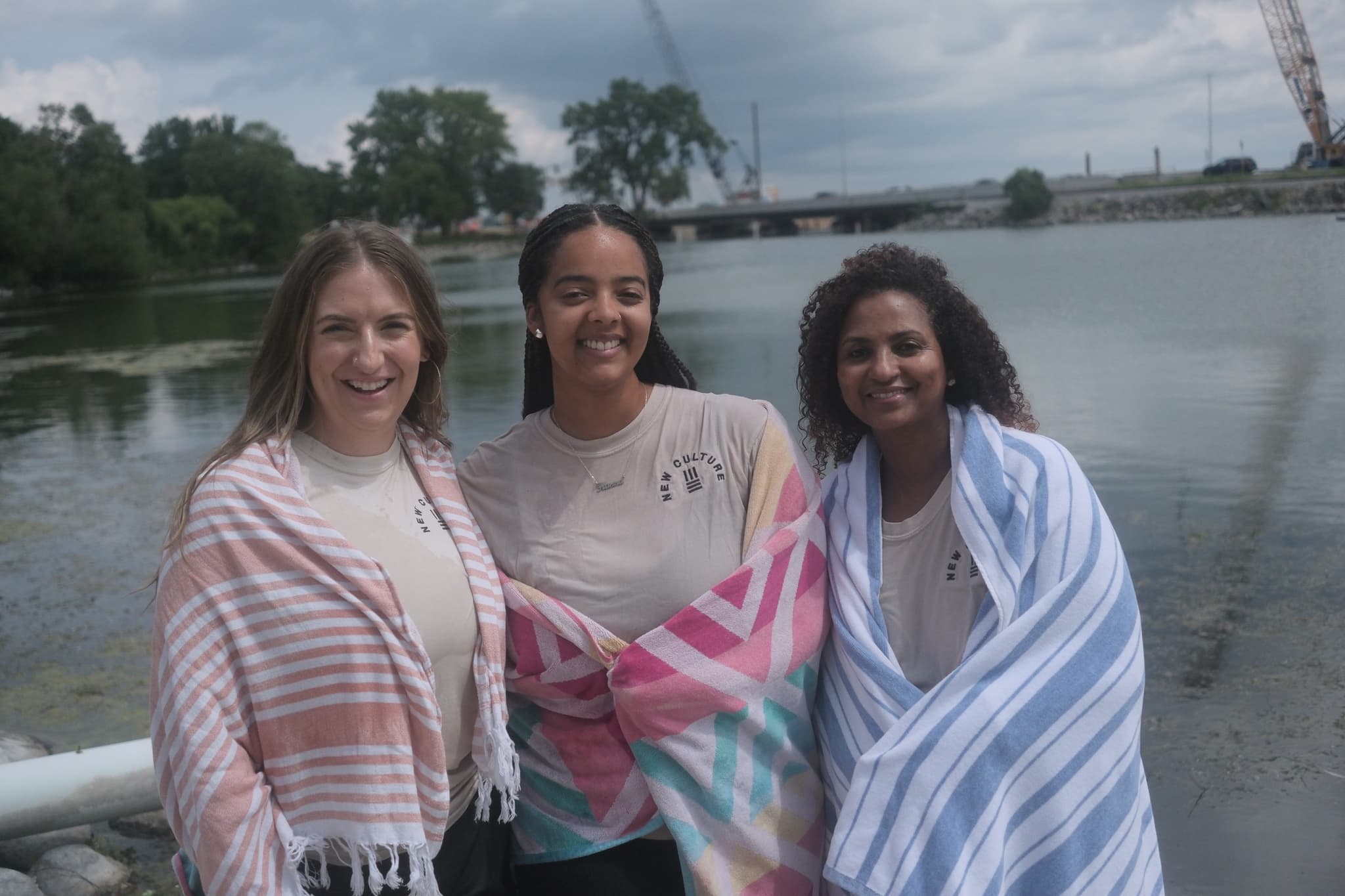 Three women from New Culture Church wrapped in towels and smiling beside Monona Bay after being baptized in Madison, Wisconsin